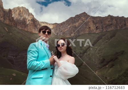 wedding couple in sunglasses and wedding suits stands stylishly against the background of high rocky mountains covered with grass. wedding couple in sunglasses and wedding suits stands stylishly against the background of high rocky mountains covered with grass. 116712683