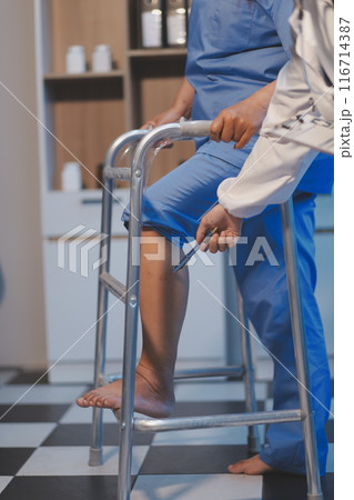 Asian young professional successful friendly female nurse in blue hospital uniform helping supporting physical therapy senior old pensioner unhealthy injury woman patient walking via four legs walker. Asian young professional successful friendly female nurse in blue hospital uniform helping supporting physical therapy senior old pensioner unhealthy injury woman patient walking via four legs walker. 116714387