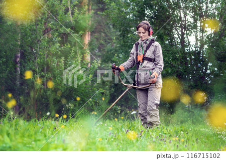 A female gardener mows the grass with a lawn mower trimmer in a clearing near the forest in the garden, doing landscaping outdoors 116715102