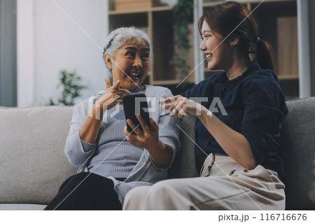 Cheerful mother and pretty teenager kid relaxing at cozy home, using mobile phone for Internet communication, resting on sofa with digital gadget, enjoying leisure, looking at camera 116716676