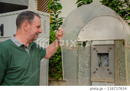 A man in a green polo shirt stands by an old, disused payphone, peering at its empty frame and exposed wires. The backdrop features urban greenery and electrical boxes. 116717634