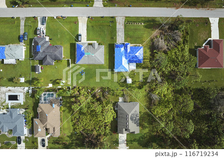 Aerial view of damaged in hurricane Ian house roofs covered with blue protective tarp against rain water leaking until replacement of asphalt shingles Aerial view of damaged in hurricane Ian house roofs covered with blue protective tarp against rain water leaking until replacement of asphalt shingles 116719234