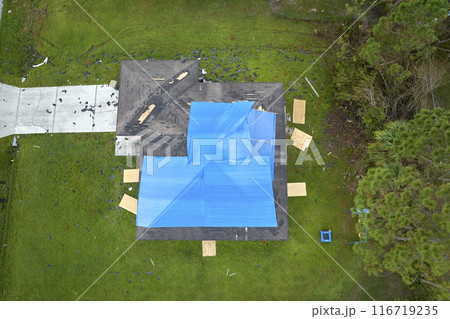 Aerial view of damaged in hurricane Ian house roof covered with blue protective tarp against rain water leaking until replacement of asphalt shingles Aerial view of damaged in hurricane Ian house roof covered with blue protective tarp against rain water leaking until replacement of asphalt shingles 116719235