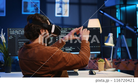 Remote software developer using VR goggles in personal office at home to do javascript programming. IT engineer wearing virtual reality headset in order to visualize lines of code, camera A Remote software developer using VR goggles in personal office at home to do javascript programming. IT engineer wearing virtual reality headset in order to visualize lines of code, camera A 116720421