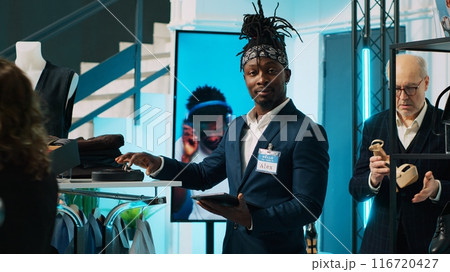 African american worker checking inventory for new clothing line placed on hangers for sale, looking at merchandise on tablet and in store. Retail assistant verifying discount list at mall. Camera A. 116720427