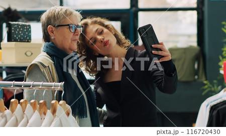 Elderly client takes pictures with her niece in clothing store, having fun while they are doing a shopping spree at the mall. Young and senior women taking photos, buying clothing items. Camera A. 116720430