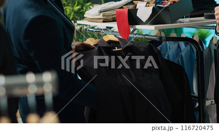 African american worker showing a black shirt and red tie to client, assisting him to buy the perfect outfit for an elegant occasion. Professional stylist gives advice to buyer at store. Camera A. 116720475