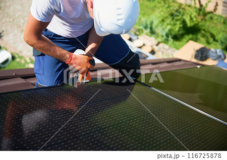 Worker building photovoltaic solar panel system on rooftop of house. Close up of man engineer in helmets and gloves installing solar module with help of hex key outdoors. Alternative, renewable energy Worker building photovoltaic solar panel system on rooftop of house. Close up of man engineer in helmets and gloves installing solar module with help of hex key outdoors. Alternative, renewable energy 116725878