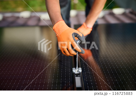 Worker building photovoltaic solar panel system on rooftop of house. Close up of man engineer in gloves installing solar module with help of hex key outdoors. Alternative and renewable energy. 116725883
