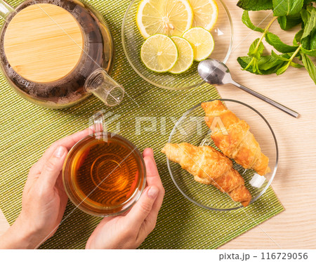 Hands holding cup of tea over a table with a glass teapot, croissants, lemon and lime slices and fresh mint on a green cutlery stand 116729056