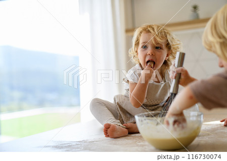 Little cute girl sitting on kitchen countertop, tasting pancake batter with finger. Young nuclear family spending weekend day indoors, cooking together. Little cute girl sitting on kitchen countertop, tasting pancake batter with finger. Young nuclear family spending weekend day indoors, cooking together. 116730074