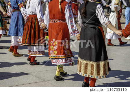 Women in bright colorful traditional Bulgarian costumes perform a horo round dance in city square 116735119