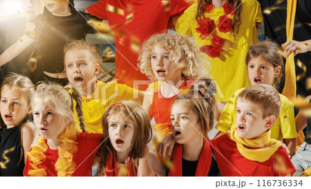 Group of children dressed in vibrant red, yellow, and black outfits spellbound watching their favorite team play on field, and golden confetti falling around them. 116736334