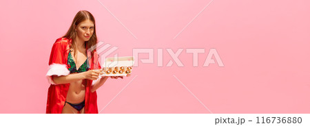 Young woman in festive lingerie and red robe holding carton egg box of golden Christmas ornaments against pink studio background Young woman in festive lingerie and red robe holding carton egg box of golden Christmas ornaments against pink studio background 116736880