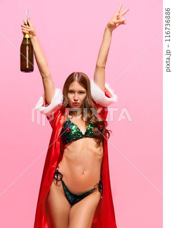 Young woman with slim body, wearing red festive gown and green bikini, standing with champagne bottle and celebrating holiday against pink studio background 116736886