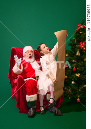 Surprised Santa sitting with girl, child reading long wish list beside beautifully decorated Christmas tree, against green studio background Surprised Santa sitting with girl, child reading long wish list beside beautifully decorated Christmas tree, against green studio background 116736988