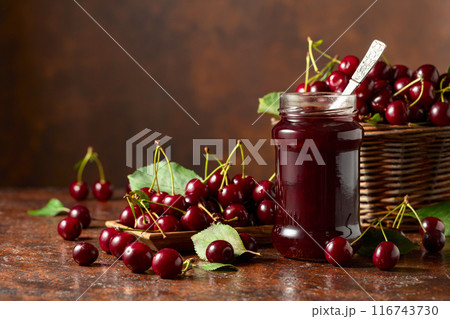 Cherry jam and fresh berries with leaves on an old brown table. Cherry jam and fresh berries with leaves on an old brown table. 116743730