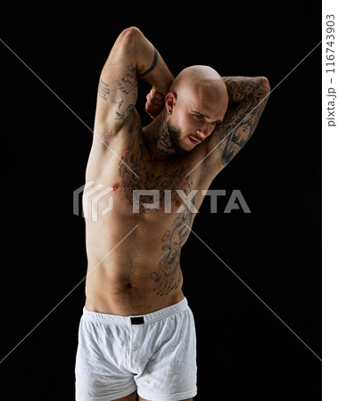 Handsome young guy with fit, relieve, muscular body with tattoos, posing shirtless in white boxers against black studio background Handsome young guy with fit, relieve, muscular body with tattoos, posing shirtless in white boxers against black studio background 116743903