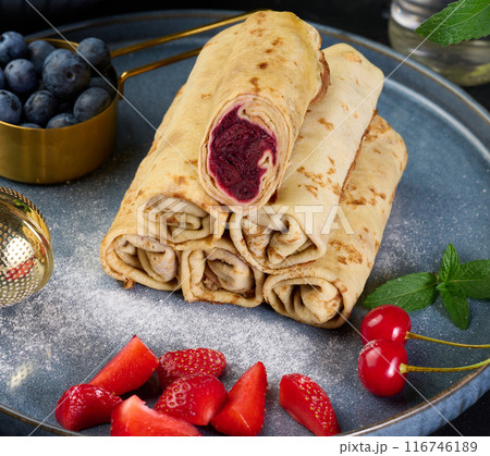 Crepes stacked in a pile, one of which is cut and filled with berry filling. The plate is also decorated with blueberries, strawberries 116746189