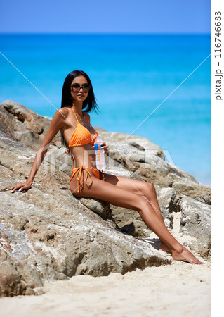 Beautiful asian girl in a orange bikini posing on a beach with rocks Beautiful asian girl in a orange bikini posing on a beach with rocks 116746683