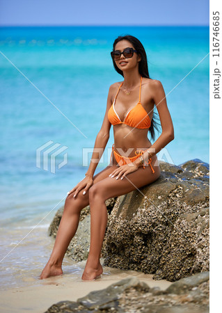 Beautiful asian girl in a orange bikini posing on a beach with rocks Beautiful asian girl in a orange bikini posing on a beach with rocks 116746685