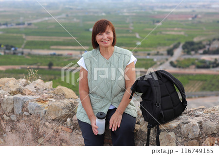 Active middle aged woman with backpack relaxes sits with water bottle on hill, looks at beautiful view after hiking up. Healthy lifestyle in retirement. Adventure seeks woman treks enjoys freedom 116749185