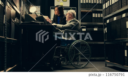 Inspector consulting with criminal expert on archived materials and autopsy records, examining folders and old cases to find connections. Private investigation in disability friendly space. Camera B. 116750791