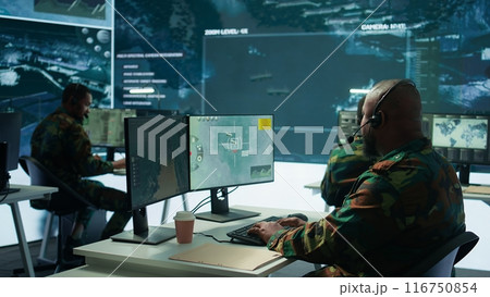 Military personnel in a base of operations using advanced computers and big screen, monitoring satellite systems to enhance security measures and process data. High tech army gear. Camera B. 116750854