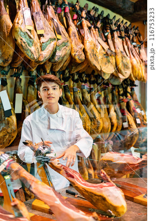 Young guy seller in white jacket selling spanish jamon in butcher shop Young guy seller in white jacket selling spanish jamon in butcher shop 116753233