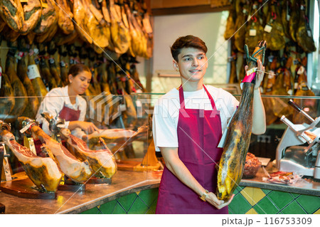 Cheerful young butcher shop salesman holding leg of Iberian ham 116753309