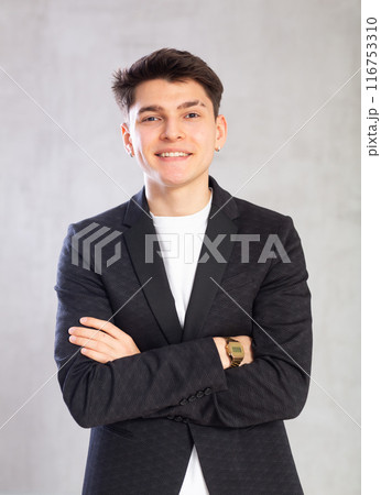 Positive male student in black jacket poses with folded arms on chest on gray background 116753310