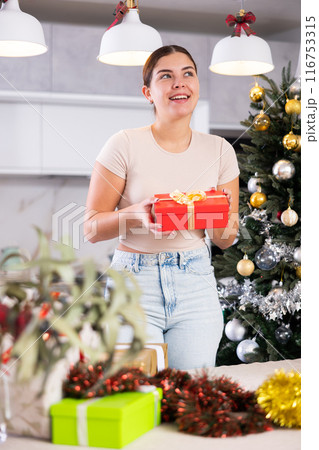 Emotional young European woman holding and enjoying wrapped gift next to Christmas tree before holidays at flat 116753315