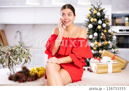 Emotional young female in red dress talking on mobile phone close to packed gift boxes and decorated Christmas tree at apartment Emotional young female in red dress talking on mobile phone close to packed gift boxes and decorated Christmas tree at apartment 116753355