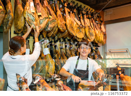 Young salesman in apron slicing Iberian jamon on jamonera in butcher shop 116753372
