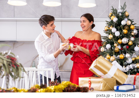 Excited young couple enjoying decorating Christmas tree together while standing in front of wrapped presents in living room 116753452