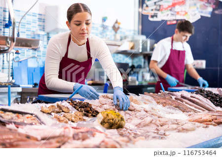 Portrait of female fishmonger showing fresh fish to woman behind counter of seafood store Portrait of female fishmonger showing fresh fish to woman behind counter of seafood store 116753464