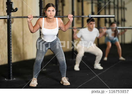 Girl in activewear exercising with barbell during group workout in gym center Girl in activewear exercising with barbell during group workout in gym center 116753857