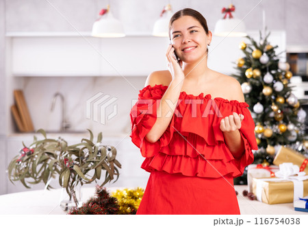 Emotional young female in red dress talking on mobile phone close to packed gift boxes and decorated Christmas tree at apartment 116754038