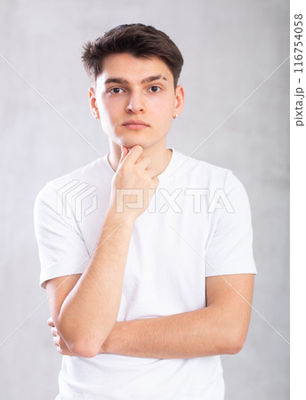 Thoughtful young man standing against background of grey unicolored wall Thoughtful young man standing against background of grey unicolored wall 116754058