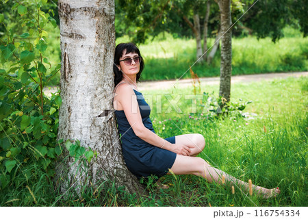 50 years old russian woman in short dress and sunglasses sitting and resting under tree shadow on the summer day  50 years old russian woman in short dress and sunglasses sitting and resting under tree shadow on the summer day  116754334