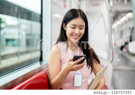 A beautiful Asian female office worker taking a sky train to work, reading text on her smartphone. 116757345