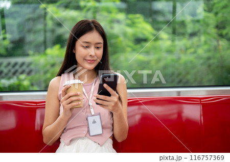 A charming Asian businesswoman is sipping coffee and reading messaged on her phone on the sky train. 116757369