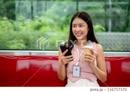 A charming Asian businesswoman is sipping coffee and reading messaged on her phone on the sky train. 116757370