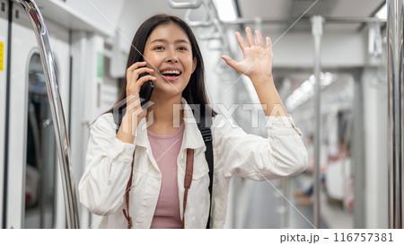 A cheerful Asian female is talking on the phone and greeting someone while commuting by sky train. 116757381