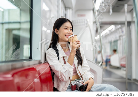 A happy young Asian female tourist is sipping coffee while sitting on a seat in a sky train. 116757388
