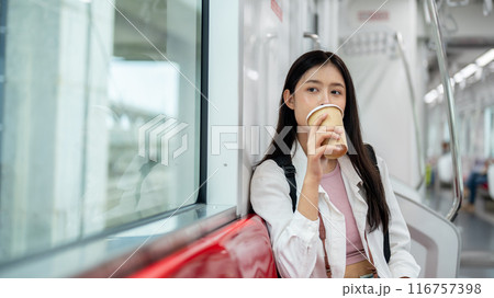 A happy young Asian female tourist is sipping coffee while sitting on a seat in a sky train. 116757398