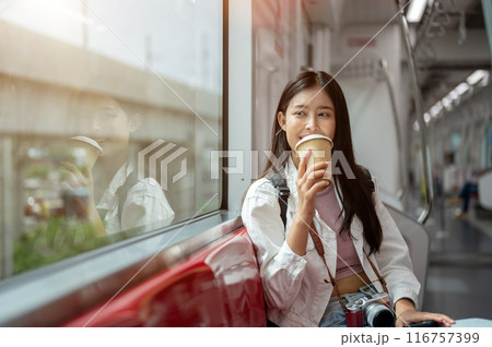 A happy young Asian female tourist is sipping coffee while sitting on a seat in a sky train. A happy young Asian female tourist is sipping coffee while sitting on a seat in a sky train. 116757399