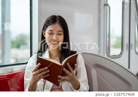 A happy Asian woman with a backpack is enjoying reading a book while taking a sky train somewhere. 116757409