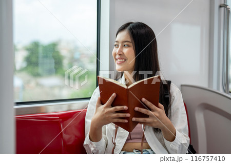 A happy Asian woman is looking at the view outside and reading a book while taking a sky train. 116757410