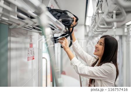 A happy female is putting her backpack in an overhead luggage space while commuting on a sky train. 116757416